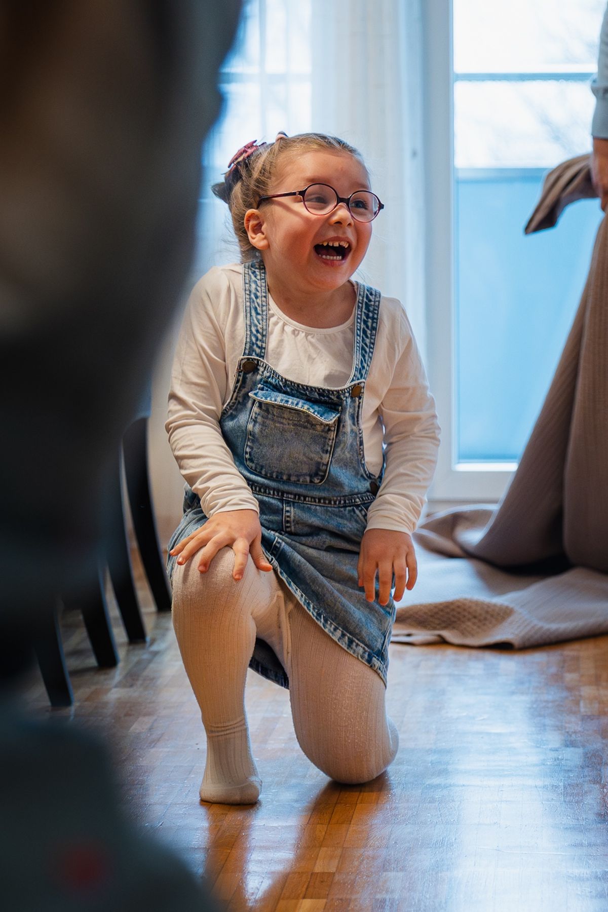 Une petite fille à lunettes rit aux éclats, à genoux sur le parquet, pendant que son papa joue avec une couverture